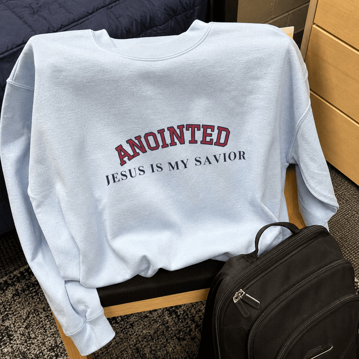 Light blue My Jesus sweatshirt draped over a chair beside a backpack